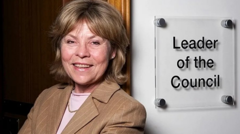 Council leader Izzi Seccombe wearing a brown pinstripe suit next to a sign which says Leader of the Council