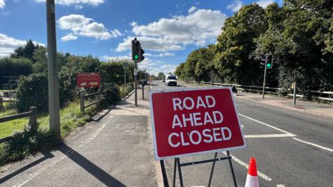 A sign reading Road Ahead Closed at Donnington Bridge in Oxford, picture taken on a sunny day. A white van is seen travelling on the road.