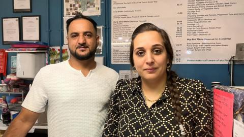 A man in a white t-shirt stand next to a woman with dark plaited hair, wearing a black and yellow patterned shirt. They stand behind the counter of a cafe, which has a large menu up on a board on the wall behind them.