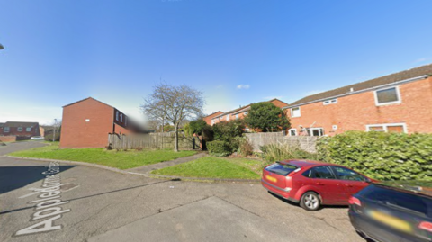 Residential street with red-brick houses, a small grassy area and tree, wooden fencing, and two parked cars, a red hatchback and a dark car, under clear blue sky.