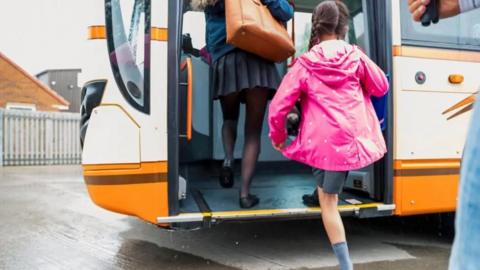 A girl with long dark hair in pig tails in a pink anorak, grey skirt and grey socks stepping up on to an orange and white bus. There is a person in front of her wearing a blue top and grey skirt and carrying a brown bag. There are signs of recent rain and a grey railing is visible in the background.