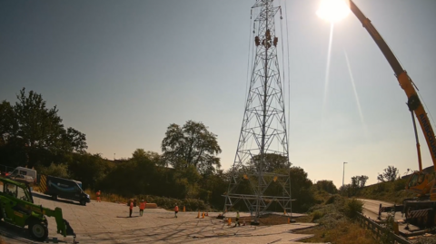 Construction site with a tall steel lattice transmission tower being erected on a prepared gravel surface. A large mobile crane on the right is supporting the tower with lifting cables attached near the top. Several workers in high-visibility orange clothing are positioned around the base of the structure. A green telehandler and parked vans are visible on the left, while trees, vegetation, and a roadside barrier line the background. Bright sunlight in the upper-right corner creates strong glare and long shadows under a clear blue sky. There are also workers on the tower.