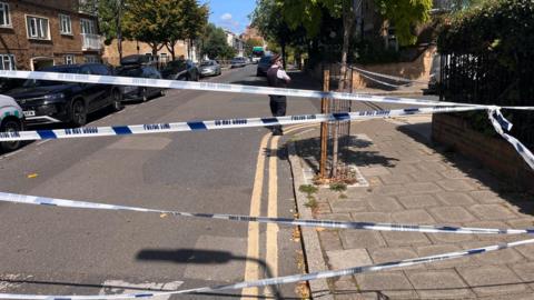 A suburban tree lined street criss-crossed with police tape. A police officer is standing beyond the tape.