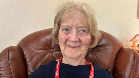 A close-up photo of 77-year-old Maureen Treharn. She has blond bobbed hair, and is wearing a red volunteer lanyard and blue jumper. She is sitting in a brown leather high back armchair and smiling at the camera. Only her head and shoulders are visible.