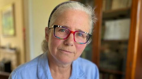 Heather is wearing a blue blouse and brightly-framed glasses. Her hair is tied back from her face and she has a neutral expression. She is in her living room with bookshelves and a television screen in the background, out of focus.