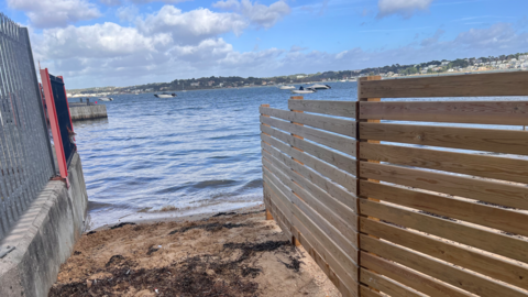 The end of the footpath where it meets Poole Harbour. On the left is a concrete wall topped with a metal fence. On the right is a wooden fence made with horizontal panels.