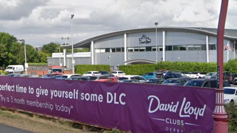 A street view image of David Lloyd gym in Derby with a purple sign in front of a car park