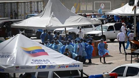 A group of staff officers in blue protective wear stand beneath a white tent