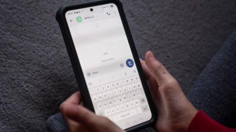 A caller holds a mobile phone in their hand. They have an entry on their screen for Jenny G and they are messaging their contact.