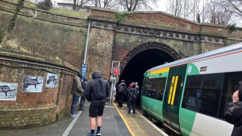 A group of people peering at a tunnel. A train is standing by at a platform.