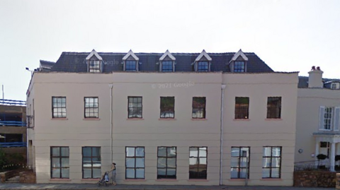 A street-view of Bermuda House on Green Street on St Helier. The building is white and has five windows at the top, seven on the second floor and seven larger windows on the ground floor. There is a woman pushing a pushchair in front of the building on the left. 