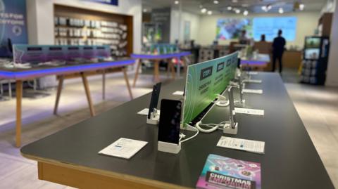 A modern shop floor of a technology/phone store, with tables display rows of mobile phones. 