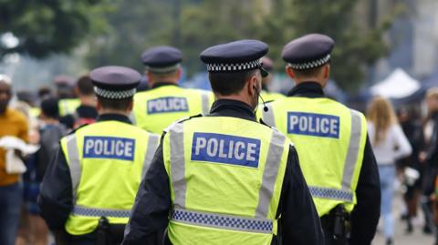 Four uniformed police officers, wearing yellow hi-vis vests and black hats with a black and white checked band, patrol at Notting Hill Carnival on 27 August 2023