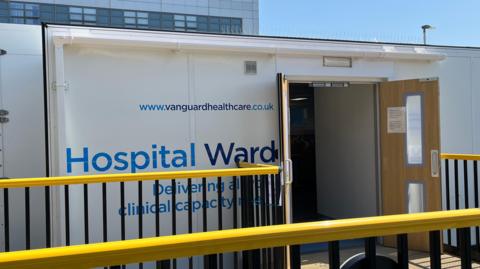 Large white rectangle building with open wooden doors and Hospital Ward written on the side in blue writing. Behind is the backdrop of the top of the hospital with glass windows and blue sky. In front of the temporary building is a yellow railing.