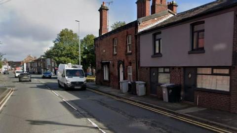 A road through Macclesfield, with a row of terrace homes seen to one side with bins out on the pavement on a sunny day. A white van is driving along one side of the road while traffic can be seen in the distance. 