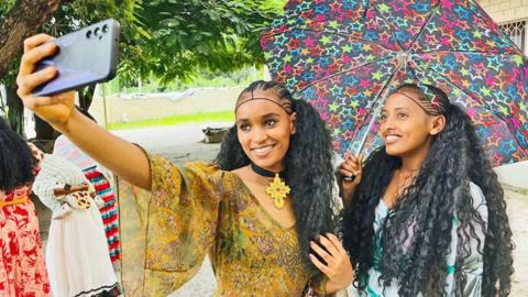 Two girls wearing colourful patterned dresses with a colourful umbrella smile and pose whilst taking a selfie. Their hair is in braids. They are outside and there are some women in dresses in the background as well as trees - Mekelle, Ethiopia. Sunday 24 August 2025.