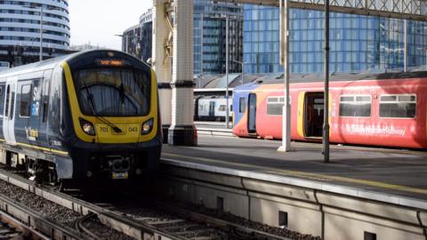 A red and yellow train with South West Railway branding at a station with another behind it