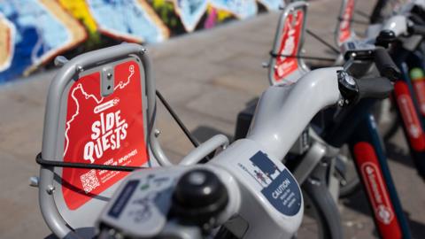 A close up shot of Santander Cycles that have red Side Quests marketing on them. 