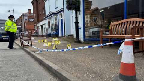 Police office stands near an area outside a shop which has police tape between some shops. Police black and yellow numbers tiles are on the pavement. 