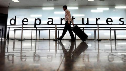 A man walking along through departures at Glasgow airport, carrying a wheeled trolley bag.