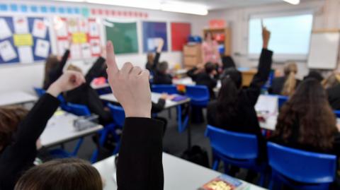 School pupils are in a classroom. Some have their hands up. 