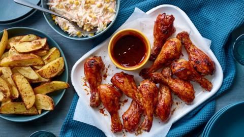 A plate of chicken wings with some sauce next to a bowls of potato wedges and bowl of coleslaw.