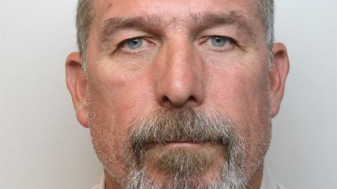 A cropped police mugshot of a man with a light grey moustache and beard. He has grey/blue eyes and is staring directly into the camera. 