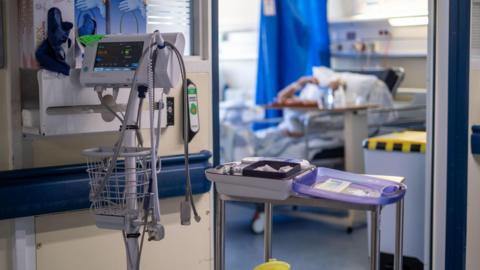 Medical equipment stands in front of a hospital room with a patient out of focus inside the room