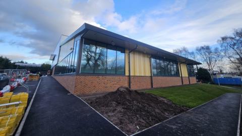 A rectangular building which has red brick at the bottom then dark grey windows and light wooden slats. The front of the building is not finished, there is a patch of grass on the right and a pile of soil on the left. The perimeter is still fenced off as construction work is ongoing. The sky is blue and cloudy in the background. 