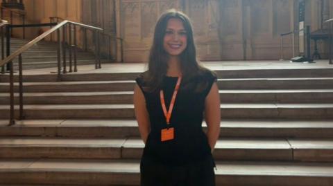 A young woman with brown shoulder length hair wearing a black outfit and orange lanyard. She is standing in front of some steps in the House of Commons