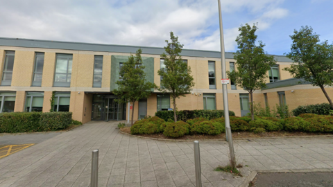 The entrance to a modern brick building with grey windows. It has a revolving door. There are bushes and trees in the mid-ground, with a path and bollards in the foreground.