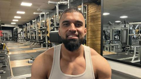 A man with short dark hair and a beard in a white vest top with rows of gym equipment behind him