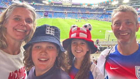 A family selfie taken inside a football stadium with the pitch and stands behind them. Amy has blonde hair and is wearing a white T-shirt, Arlo has shoulder-length blonde hair and is wearing an England bucket hat, Flo has long blonde hair and is wearing a red England hat, and Neil has short hair and is wearing an England shirt. They are all smiling at the camera.