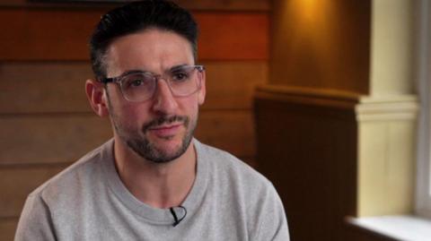 Yoni Finlay stares forward while wearing a grey sweatshirt. He has black hair and is wearing transparent glasses. 