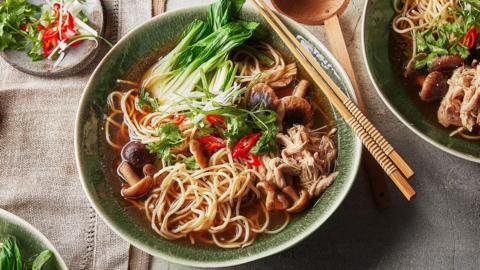 A bowl of chicken noodle soup with mushrooms and pak choi, a pair of wooden chopsticks is resting on top of the bowl.