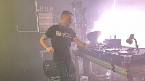A man with short hair, a black shirt and jeans, with headphones around his neck, stands next to DJ decks.