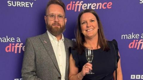 Barry Ahearn and his wife Naomi at the Make a Difference Awards 2025. Barry is wearing a grey suit jacket and navy trousers. He has glasses and a beard. Natalie is wearing a navy jumpsuit and has her hair down. Both are holding champagne glasses and are stood in front of a purple 'Make a Difference' banner. 