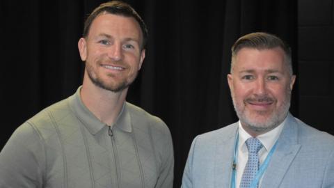 Tommy Smith stands on the left of the picture wearing a light green top with a zip and collar. Alan is on the right wearing a light blue suit and tie. Both are smiling.