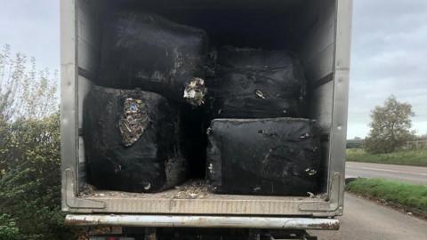 The back of a silver-white lorry trailer, with four black bales visible. There's a split in one of the bales, and you can see macerated waste inside. It's parked in a lay-by, with trees and hedges visible on the left. On the right, you can see part of the road. The photo is taken during the day and the sky is grey and cloudy.