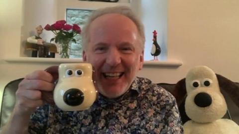 Nick Park has grey hair and is wearing a floral shirt. He is sat on a sofa with a Gromit sift toy next to him and is holding a Gromit shaped mug. On a window ledge behind is a vase of red flowers, a penguin character, and the Wallace character from his animations.