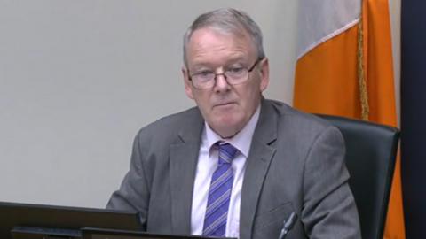 Brian Stanley is sitting in a committee. He's wearing black glasses and a grey suit with a white shirt and a purple tie. The Irish tricolour flag is behind him.