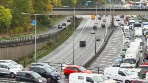 Queues of traffic on the M25 motorway near Waltham Cross in Hertfordshire. Cars are queueing on bridges going over the M25. There are trees lining the road.