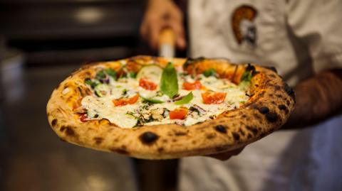 Stock photo of pizza chef presenting a pizza fresh out of the woodfired oven. The pizza is topped with cheese, tomatoes and basil. It has browned marks around its crust.