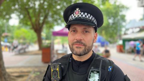 A policeman with a short beard looking at the camera with a treelined street out of focus behind him.