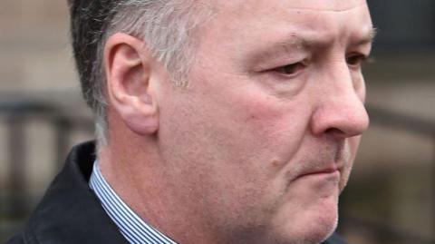 Ian Paterson head shot. He is pictured looking to the right and wearing a black jacket and stripy shirt collar. 