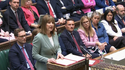 Chancellor Rachel Reeves standing at the dispatch box in he House of Commons reading from a folder, with fellow MPs behind her on green benches