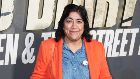 Gurinder Chadha, had long, dark hair, wearing an orange blazer and a denim shirt underneath