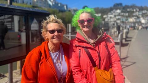 Two women stood side by side, both are wearing sunglasses and smiling. The woman on the left has short blonde hair and is wearing a purple top and red zip up hoodie. The woman on the right is wearing a red coat and has an orange bag and has short green hair. Behind them is a seaside town. 