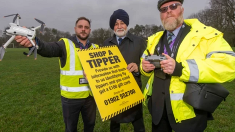 Council officers and councillor Bhupinder Gakhal stand in a field with a drone and a fly-tipping poster. The two council officers are wearing high-vis jackets over their dark trousers and jackets. Mr Gakhal is holding the yellow poster with the words 'Shop a Tipper' on and a phone number.