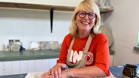 Linda smiles at the camera. She wears glasses and an orange MND association T-shirt with a beige apron over the top. She leans on a table where she has been doing pottery.